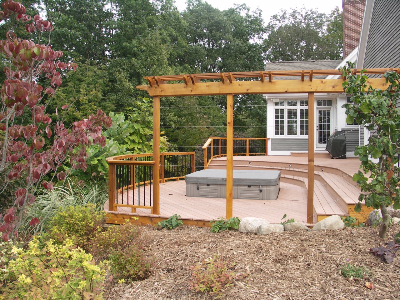 A pergola and deck setup with a curved layout design to fit a hot tub inset. For the best deck builders in Rockford MI has to offer, call Precision Decks & Sunrooms.