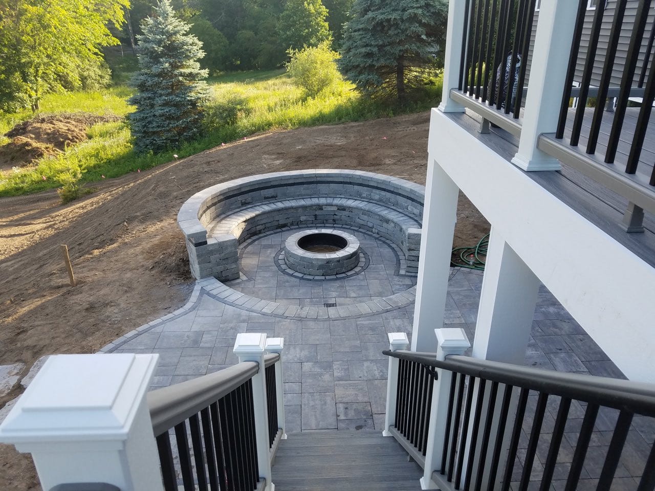 An outdoor fire feature (fire pit) being built in the middle of a circular stone patio set, steps away from the steps of an upper-floor deck.