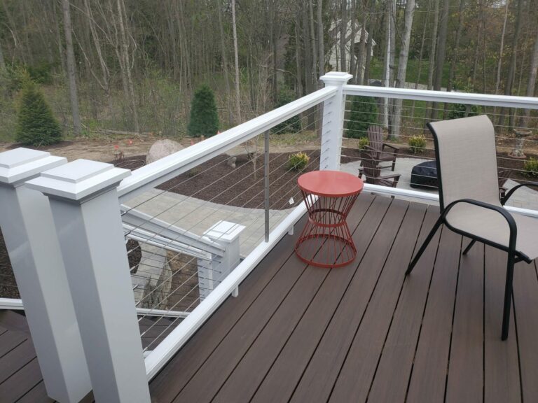 A patio and deck combo in a residential backyard. A fire pit surrounded by Adirondak chairs sits on the patio underneath.
