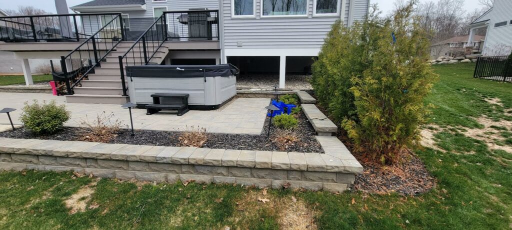 Photo of a stone paver patio with a hot tub and composite deck leading up to house.