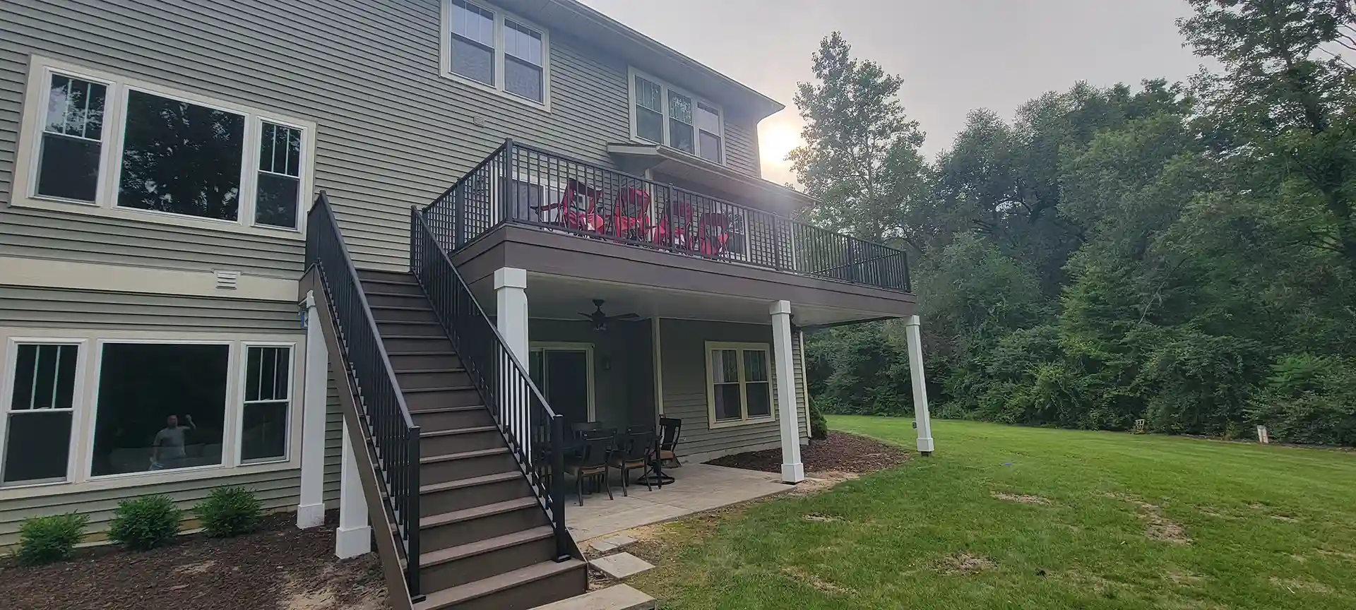 Photo of large elevated deck with black railings, red chairs, and covered patio below. 