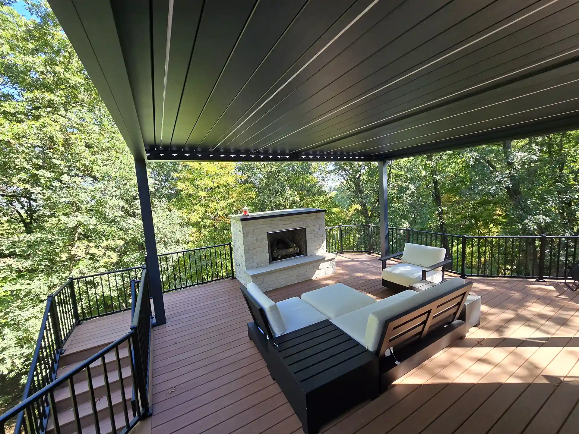 deck cost in grand rapids mi Photo of a modern outdoor deck with black railing, a louvered pergola roof, white cushioned seating, and a stone fireplace.