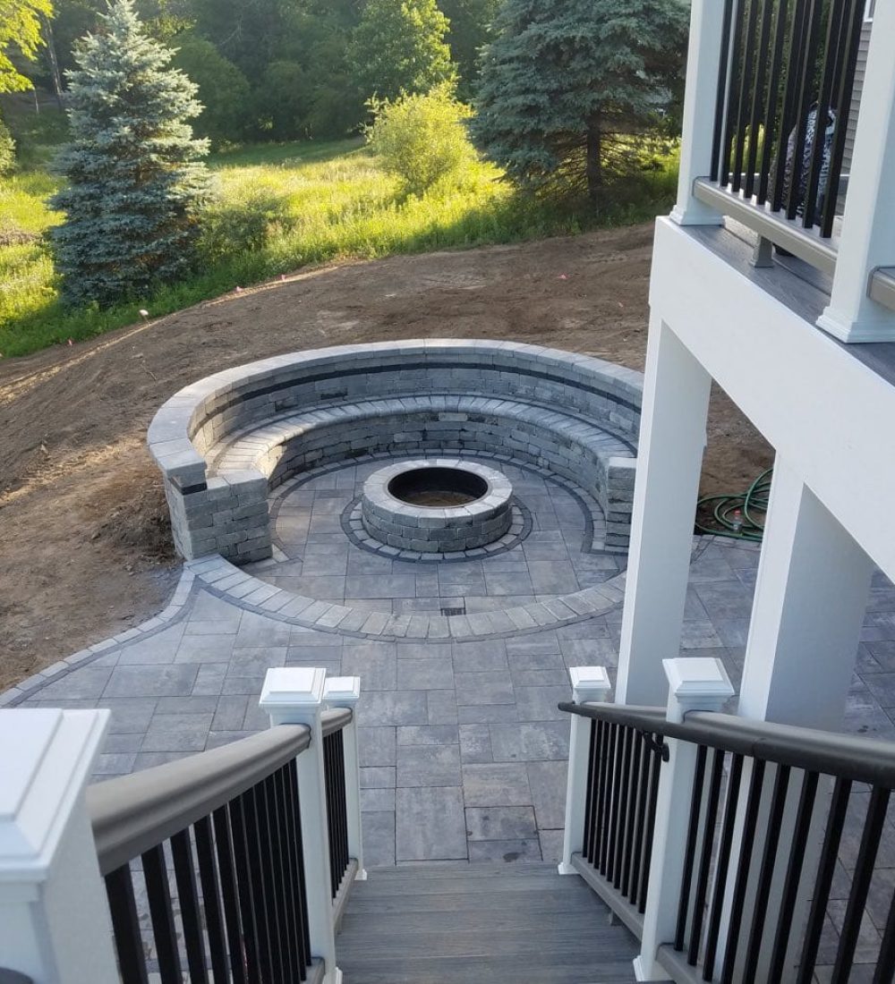 An outdoor fire feature (fire pit) being built in the middle of a circular stone patio set, steps away from the steps of an upper-floor deck.